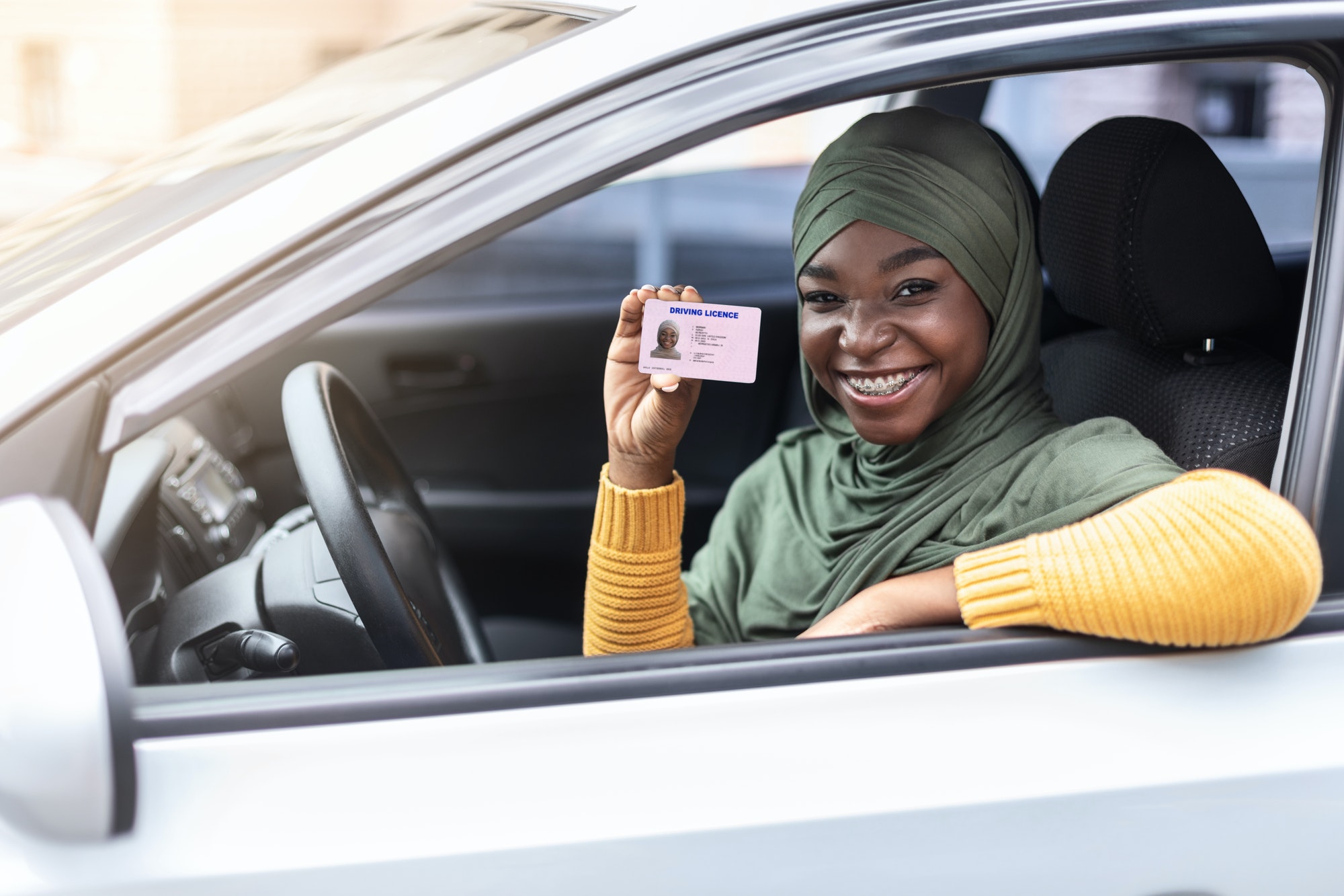 Happy black muslim woman in hijab sitting in car, showing driver license